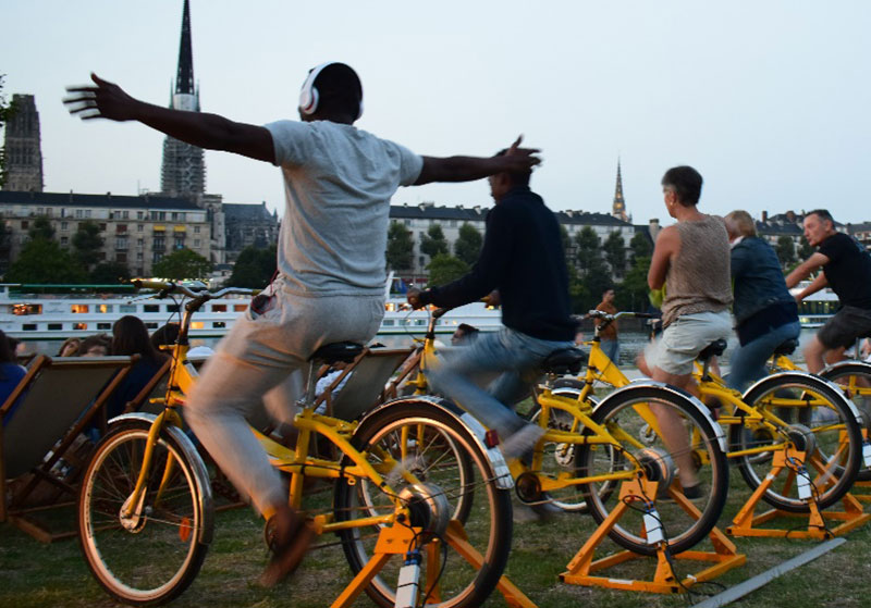 Photo de personnes qui pédalent sur des vélos jaunes face à l'écran (hors champ) d'un cinéma en plein air.